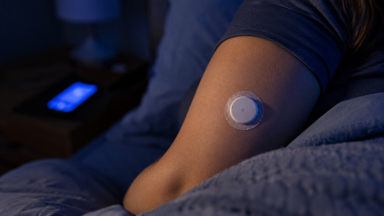 Close-up of a continuous glucose monitor sensor patch on a sleeping person's upper arm, capturing an undetected overnight low before a morning rebound
